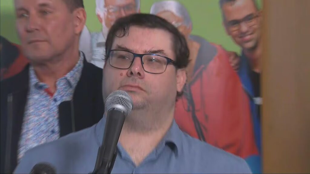 A photo of a middle aged mad wearing glasses standing at a microphone